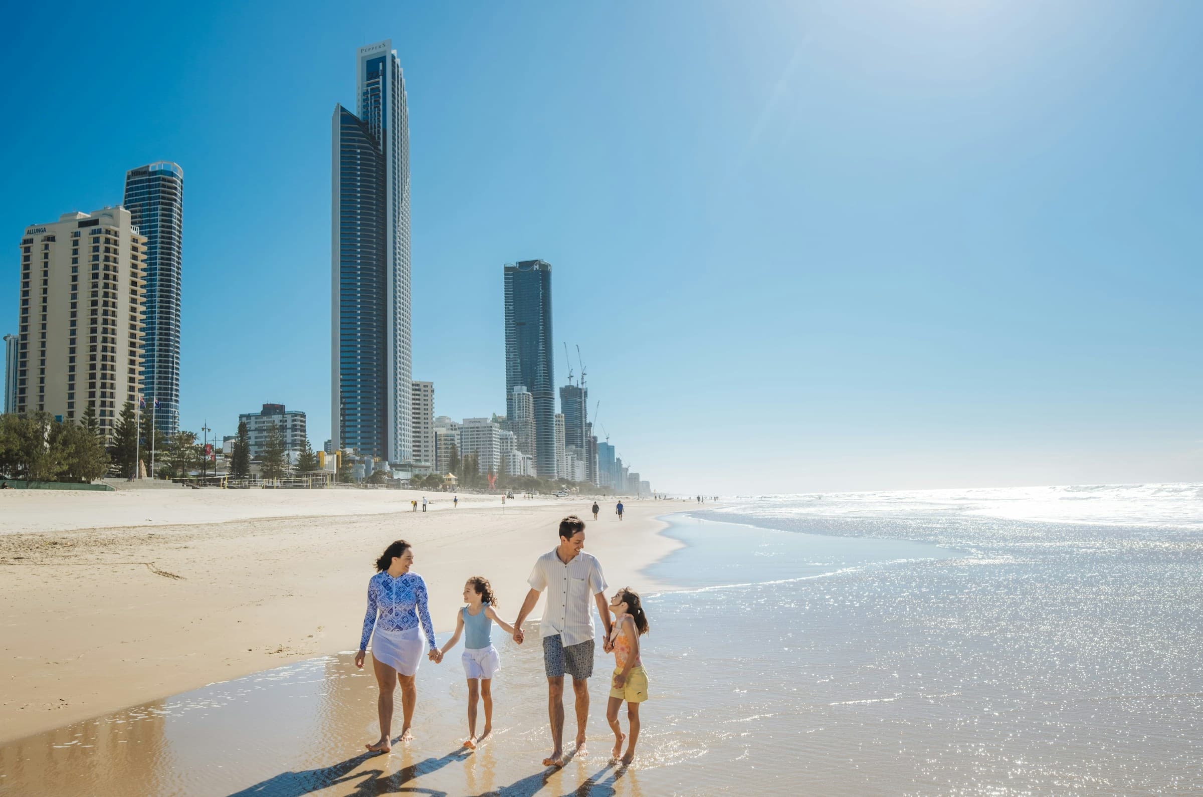A family holding hands and walking along a sunny beach with high-rise towers behind - Family Beach Holidays