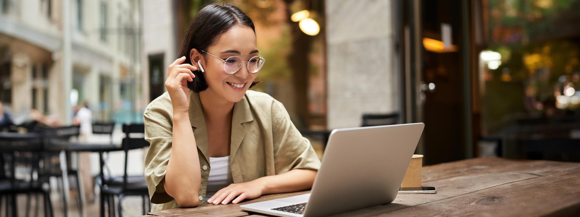 A smiling woman with an earbud video-calling on her laptop at an outdoor cafe
